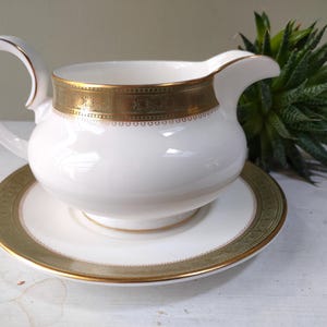 May include: White porcelain gravy boat with a gold band around the top and a matching saucer. The gravy boat has a curved handle and a spout. The gold band has a decorative pattern. The set is on a white surface.
