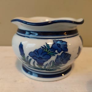 May include: A small white ceramic planter with a blue and green floral design. The planter has a scalloped rim and a blue band around the top.