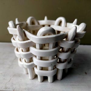 May include: A small, off-white ceramic bowl featuring a decorative woven basket design. The structure is composed of interlocking, rounded ceramic strips that create a lattice pattern, with vertical pointed elements extending along the top rim.