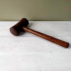 May include: A solid brown wooden mallet featuring a cylindrical head and a long, straight handle. The tool has a smooth, polished finish with visible wood grain patterns throughout the entire structure, showcasing a simple and functional design.
