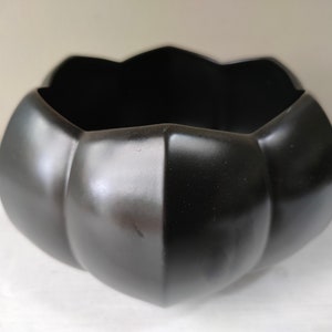 May include: A black, decorative bowl with a unique, petal-like design. The bowl has a glossy finish and a wide opening. The exterior features a series of rounded, vertical segments, creating a textured appearance. The bowl is likely made of ceramic or a similar material.