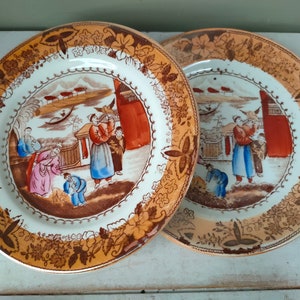 May include: Two vintage ceramic plates with a gold and brown floral border. The centre of each plate features a hand-painted scene of people in traditional Chinese clothing.