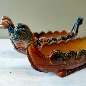 May include: Two ceramic Viking ship-shaped bowls. The bowls are brown and orange with decorative details, including dragon heads at the bow and stern. The bowls have a scalloped edge and are likely decorative or for serving.