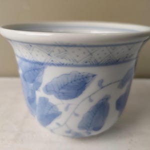 Op de afbeelding: Een witte keramische beker met een blauw bloemmotief. De beker heeft een uitlopende rand en een patroon van blauwe bladeren en wijnranken. Het bovenste gedeelte heeft een geometrisch ontwerp. De beker is waarschijnlijk voor thee of koffie.