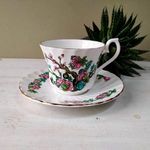 May include: White teacup and saucer set with a floral design in pink, green, and black. The teacup has a gold rim and handle, and the saucer has a matching floral border. The set is placed on a white surface.