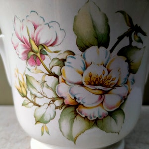 May include: White ceramic planter with a floral design featuring pink, white, and yellow roses with green leaves.