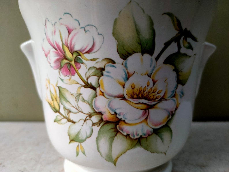 May include: White ceramic planter with a floral design featuring pink, white, and yellow roses with green leaves.