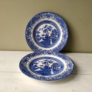 May include: Two blue and white ceramic plates with a Willow pattern design. One plate is round, and the other is oval. The plates feature a detailed landscape scene with a bridge, pagoda, and birds. The rims have a decorative border.