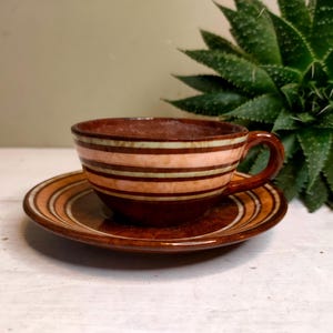 Puede incluir: Un juego de taza y platillo de cerámica. La taza y el platillo son marrones con rayas horizontales en tonos beige y crema. La taza tiene un asa. El juego está sobre una superficie blanca.