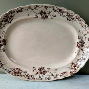 May include: Oval serving platter with a scalloped edge. The off-white ceramic platter features a brown floral and vine pattern around the rim. The platter is approximately 46 cm long.