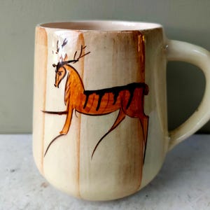 May include: A ceramic mug with a cream-colored glaze and vertical brown brushstroke stripes. The front features a hand-painted orange deer illustration with black accents. The mug has a rounded body and a curved handle on the right side.