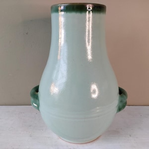 Può includere: Un vaso in ceramica verde chiaro con due manici verdi. Il vaso ha un corpo arrotondato e un collo stretto.