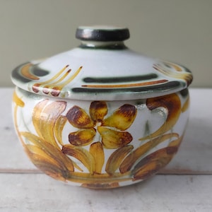 Puede incluir: Un tarro de cerámica blanca con tapa, decorado con diseños florales marrones y amarillos. El tarro tiene una banda verde y marrón alrededor de la parte superior.