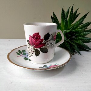 Può includere: Tazza e piattino bianchi con una rosa rosso scuro. La tazza e il piattino presentano un motivo floreale. Il piattino ha un bordo dorato. Un classico set da tè.