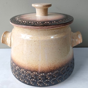 May include: A beige ceramic crock with a lid, featuring a brown and black floral design around the rim and base. The crock has two handles on the sides.