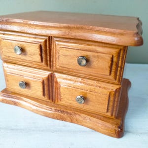 May include: A small wooden jewellery box with four drawers. The box is stained a warm, light brown colour and features silver-toned knobs. The design is classic and the box has a curved base.