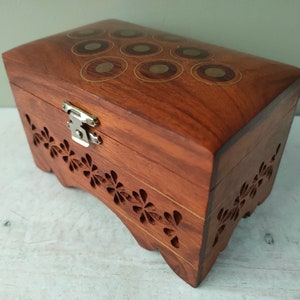 Puede incluir: Una pequeña caja de madera con tapa con bisagras y cierre de latón. La parte superior de la caja está decorada con un patrón de círculos incrustados. Los lados de la caja están tallados con un diseño floral.