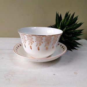 May include: A white teacup and saucer set with gold floral detailing. The teacup has a wide rim and a rounded shape, while the saucer is round with a matching gold trim. The set is placed on a white surface, with a green plant in the background.
