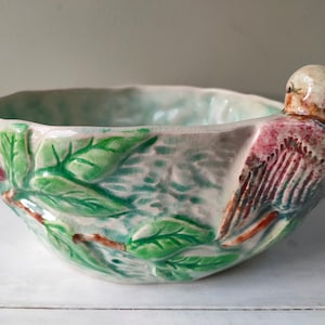 Op de afbeelding: Een witte keramische kom met een groen en bruin vogelontwerp. De kom heeft een gegolfde rand en is versierd met groene bladeren en een vogel met bruine en rode veren.