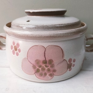 Puede incluir: Una cacerola de cerámica blanca con tapa marrón y diseño floral rosa. La cacerola tiene dos asas y un pequeño agujero en la tapa.