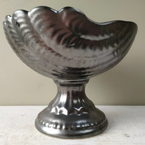 May include: A large, silver-colored ceramic bowl with a scalloped edge and a pedestal base. The bowl is shaped like a seashell.