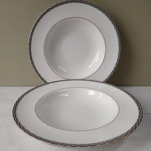 May include: Two white ceramic soup bowls with a decorative border. The border features a repeating pattern of blue, gold, and black. A thin gold line encircles the inner bowl. The bowls are on a white surface.