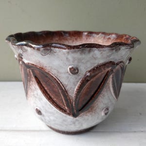 May include: A ceramic planter with a white and brown speckled finish. The planter features a scalloped rim and a repeating leaf design in dark brown. Small, raised dots accent the design.