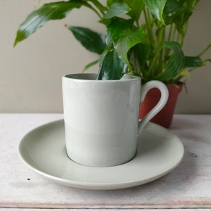 Puede incluir: Un juego de taza y platillo de cerámica verde claro. La taza tiene un diseño sencillo y un asa redondeada. El platillo es ligeramente más grande que la taza y tiene una superficie lisa.