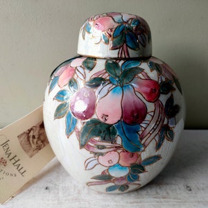 Puede incluir: Jarrón de cerámica blanca con tapa, decorado con un diseño floral con peras rosas, azules y verdes y hojas. El jarrón tiene un borde dorado y está etiquetado "Jena Hall Inspirations from the ordinary".