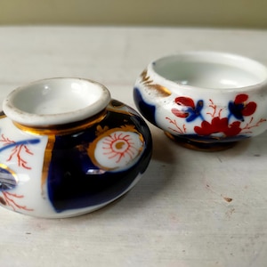 May include: Two antique porcelain pieces. One is a small, round inkwell with a narrow opening, and the other is a small, open bowl. Both are white with blue, red, and gold accents. The inkwell has a dark blue and white design with a red and white floral pattern.