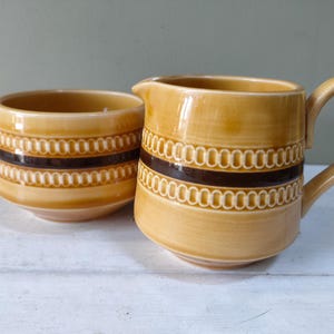 May include: A vintage ceramic creamer and sugar bowl set. The pieces are a warm yellow-brown color with a repeating pattern of white ovals and a dark brown band. The creamer has a handle and a spout.