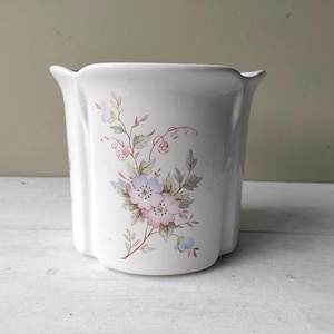Pode incluir: Um vaso de cerâmica branca com borda recortada e lados verticais canelados. A frente apresenta um delicado decalque floral com flores rosa e azuis, folhas verdes e caules castanhos finos dispostos num padrão botânico vertical.