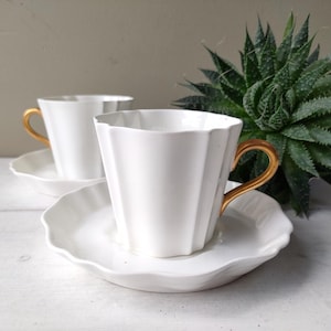 May include: Two white teacups with gold handles and matching saucers. The cups have a fluted design and sit on a white surface. A green succulent plant is in the background.