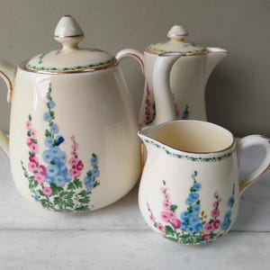 Peut inclure: Service à thé en céramique crème, comprenant une théière, un pot à lait et une verseuse. L'ensemble est orné de motifs floraux roses et bleus et de bordures dorées. La théière a un couvercle avec un bouton.