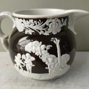 May include: A dark brown ceramic pitcher with a white interior and handle. The pitcher features white relief designs of floral patterns and classical figures. The spout is white and the pitcher has a round shape.