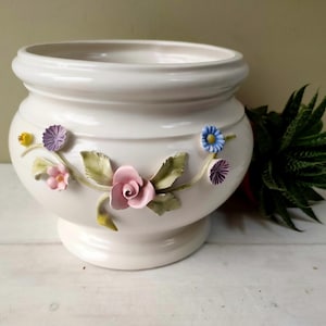 Puede incluir: Una maceta de cerámica blanca con un cuerpo ancho y redondeado y una base de pedestal. La maceta está adornada con adornos florales tridimensionales de colores, que incluyen rosas rosas, flores azules y moradas y hojas verdes. Una pequeña planta suculenta está al lado.