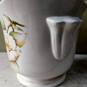 May include: White ceramic planter with a scalloped rim and a floral design featuring white flowers and green leaves. The planter has a handle on the side.