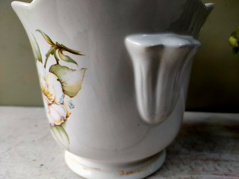May include: White ceramic planter with a scalloped rim and a floral design featuring white flowers and green leaves. The planter has a handle on the side.