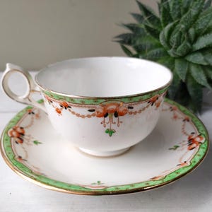 May include: White teacup and saucer set with a green and orange floral design. The teacup has a curved handle and sits on a matching saucer. The set is trimmed with gold accents, creating a vintage aesthetic.
