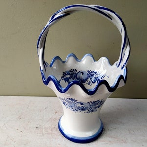 May include: A white ceramic basket with a blue and white floral design and a blue handle. The basket has a scalloped rim and a flared base.