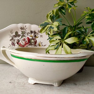 Pode incluir: Um molho de cerâmica branca com uma borda verde e dourada. O molho tem um design floral no lado e está sobre uma superfície branca.
