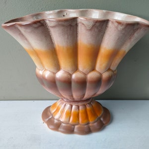 May include: A decorative ceramic vase with a fluted, fan-shaped design. The vase features a color gradient of brown, tan, and orange hues. It has a pedestal base and a scalloped rim. This vintage piece is suitable for floral arrangements or as a decorative accent.