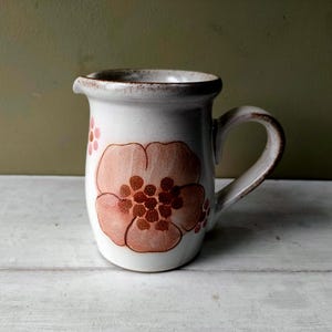 Op de afbeelding: Een witte keramische kan met een groot, gestileerd perzikbloemmotief. De kan heeft een bruine rand en handvat, en een kleine tuit. De bloem heeft een donkerbruin centrum. De kan staat op een wit oppervlak.