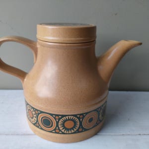 May include: A tan ceramic teapot with a lid and a curved handle. The teapot has a spout and a decorative band of orange, green, and black geometric patterns around the base. The teapot is likely vintage.