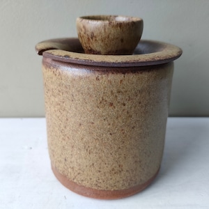 Puede incluir: Un tarro de cerámica marrón con tapa y un pequeño cuenco de cerámica marrón en la parte superior. El tarro tiene un esmalte marrón y beige moteado.