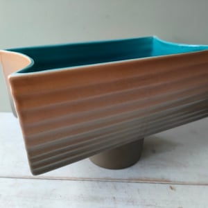 Op de afbeelding: Een rechthoekige keramische plantenbak met een tealblauwe binnenkant. De plantenbak heeft een getextureerde, bruine buitenkant met een verhoogde basis.