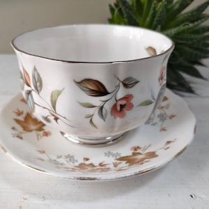 Pode incluir: Uma chávena de chá e um pires brancos com um padrão floral. A chávena tem uma borda ondulada e o pires tem uma borda dourada.