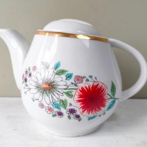 Puede incluir: Tetera de cerámica blanca con tapa y ribete dorado. La tetera está decorada con un diseño floral con una gran margarita, una flor roja y flores rosas y moradas más pequeñas con hojas verdes y azules.