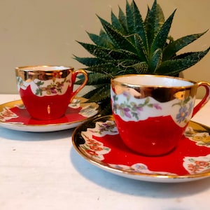 May include: Two red and gold teacups and saucers with floral designs. The cups and saucers have a gold rim and a white floral pattern. The cups are red with a gold rim and handle. The saucers are red with a gold rim and floral pattern.