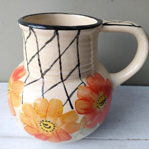 May include: Cream ceramic jug with a black rim and handle. It features orange and yellow floral designs and a black crisscross pattern. A decorative kitchen item, ideal for serving drinks or as a vase.